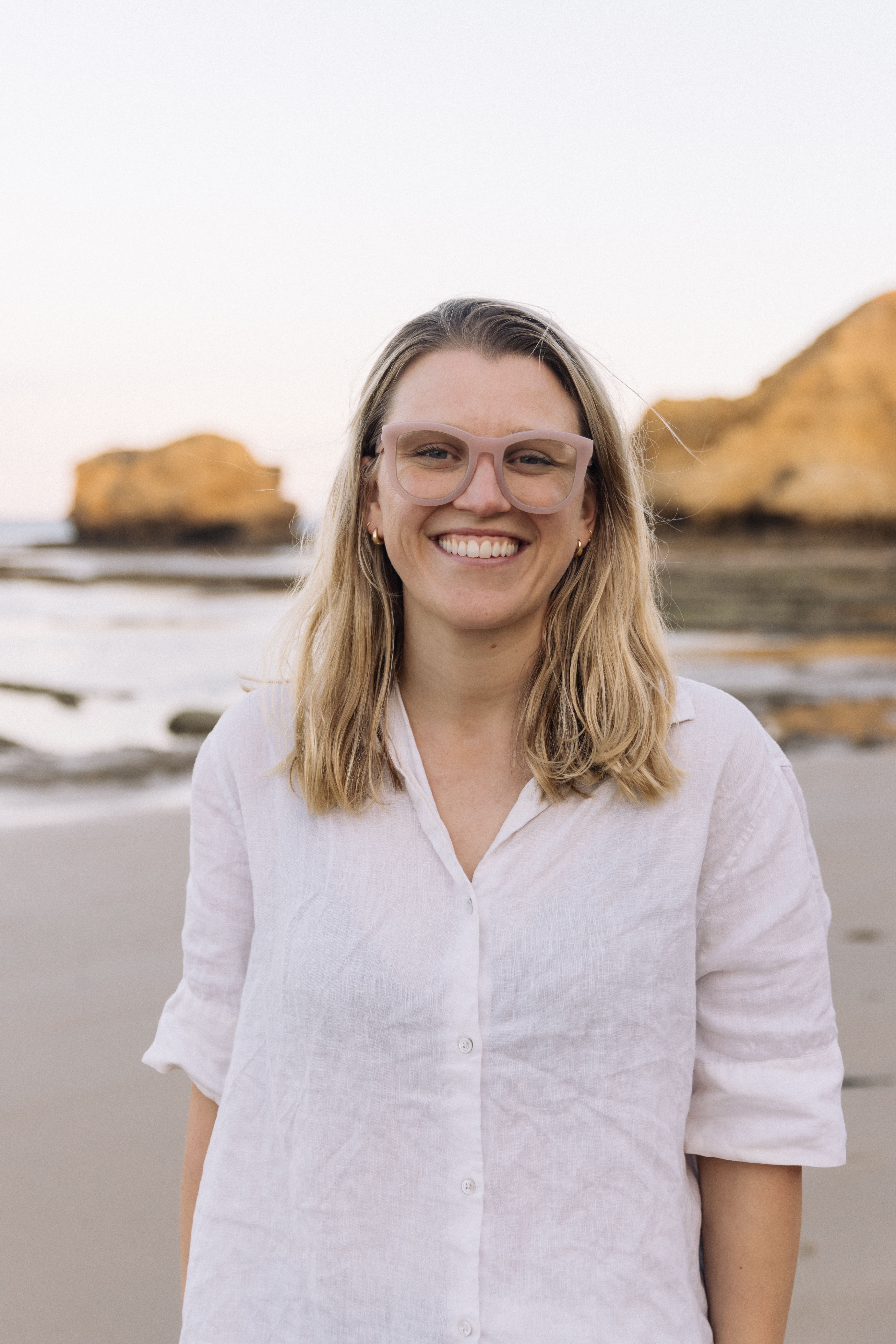 A woman named Kate is standing on a beach, smiling. She has blonde hair, wears glasses, and a white button-up shirt. The background features ocean water, rocks, and sand under soft, natural light.