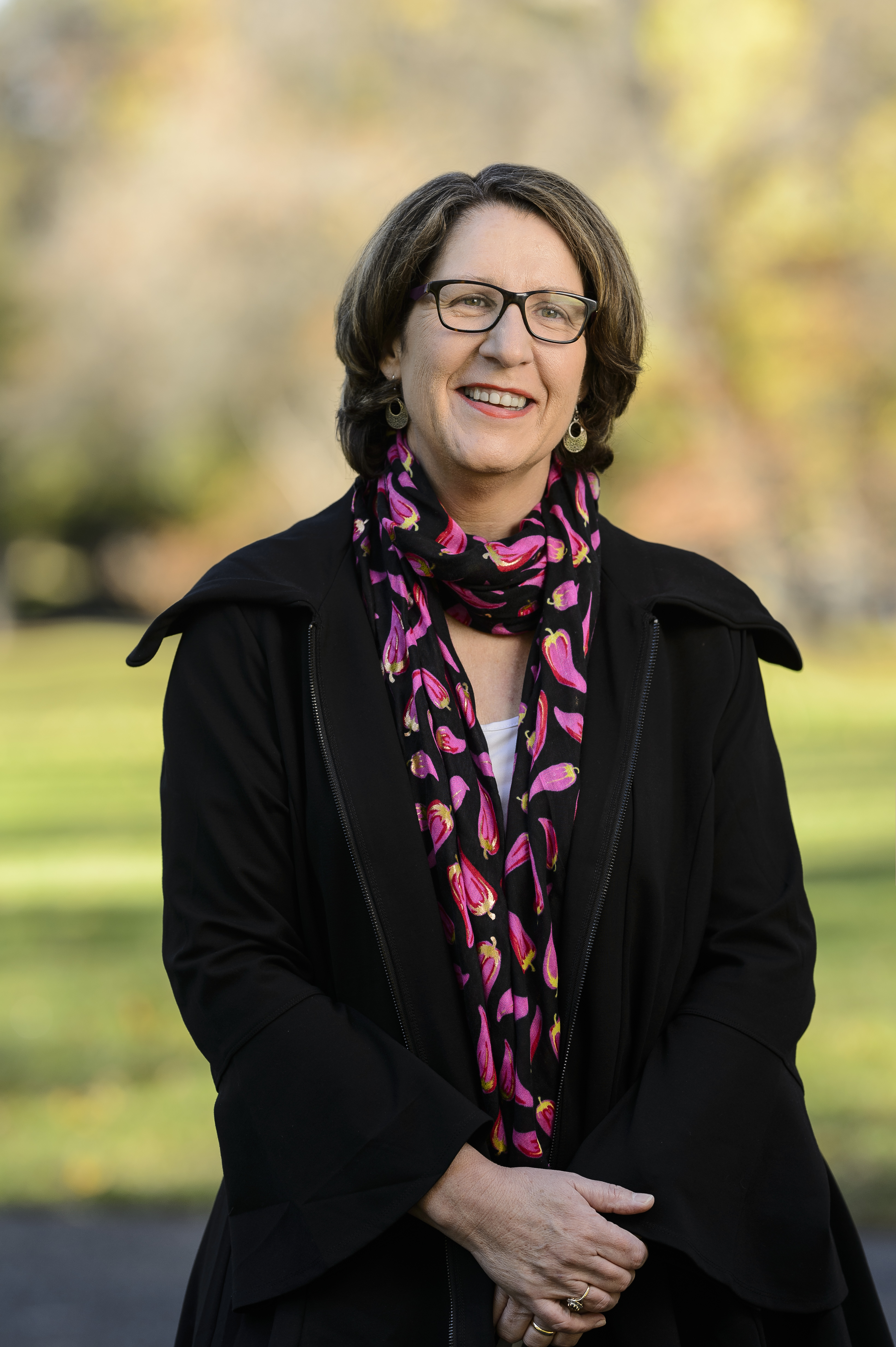 Rhonda Garad, wearing glasses and a black coat, smiles warmly. She has shoulder-length brown hair and a vibrant scarf with pink floral patterns. She stands outdoors with a softly blurred background of green grass and trees.