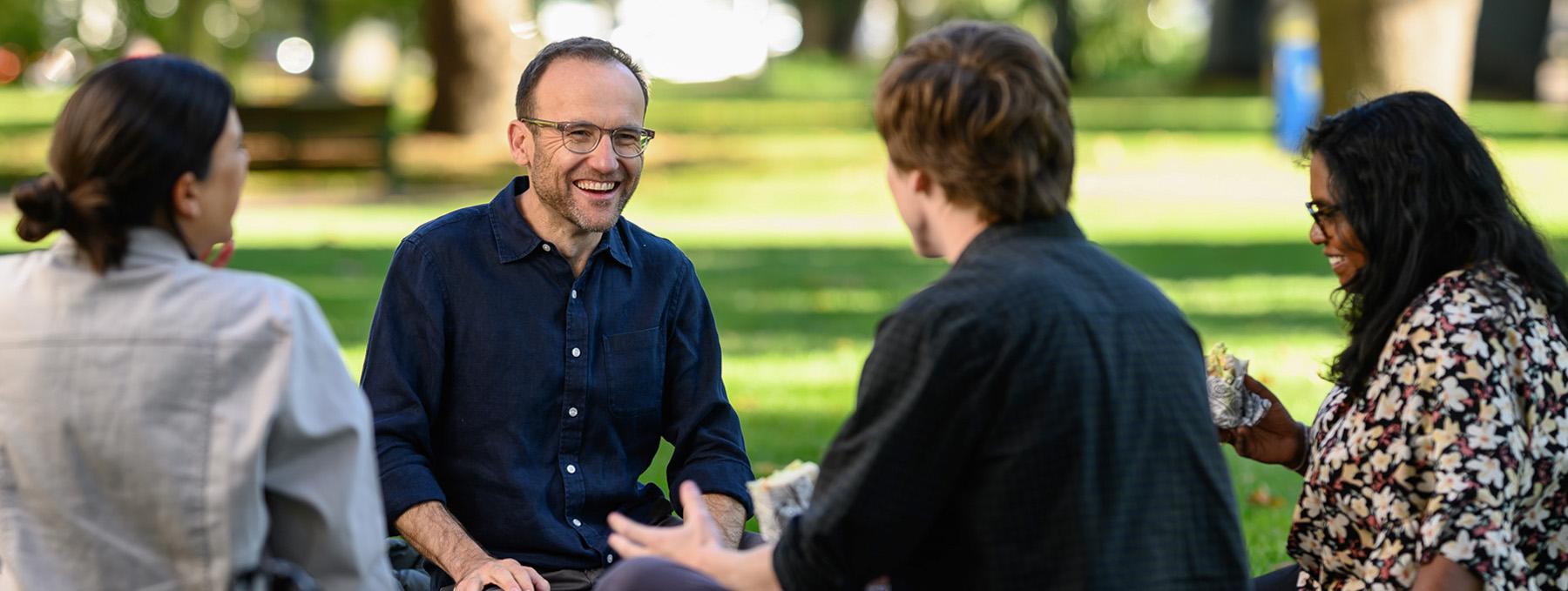 Adam Bandt smiling and sitting on the grass in a park chatting with locals